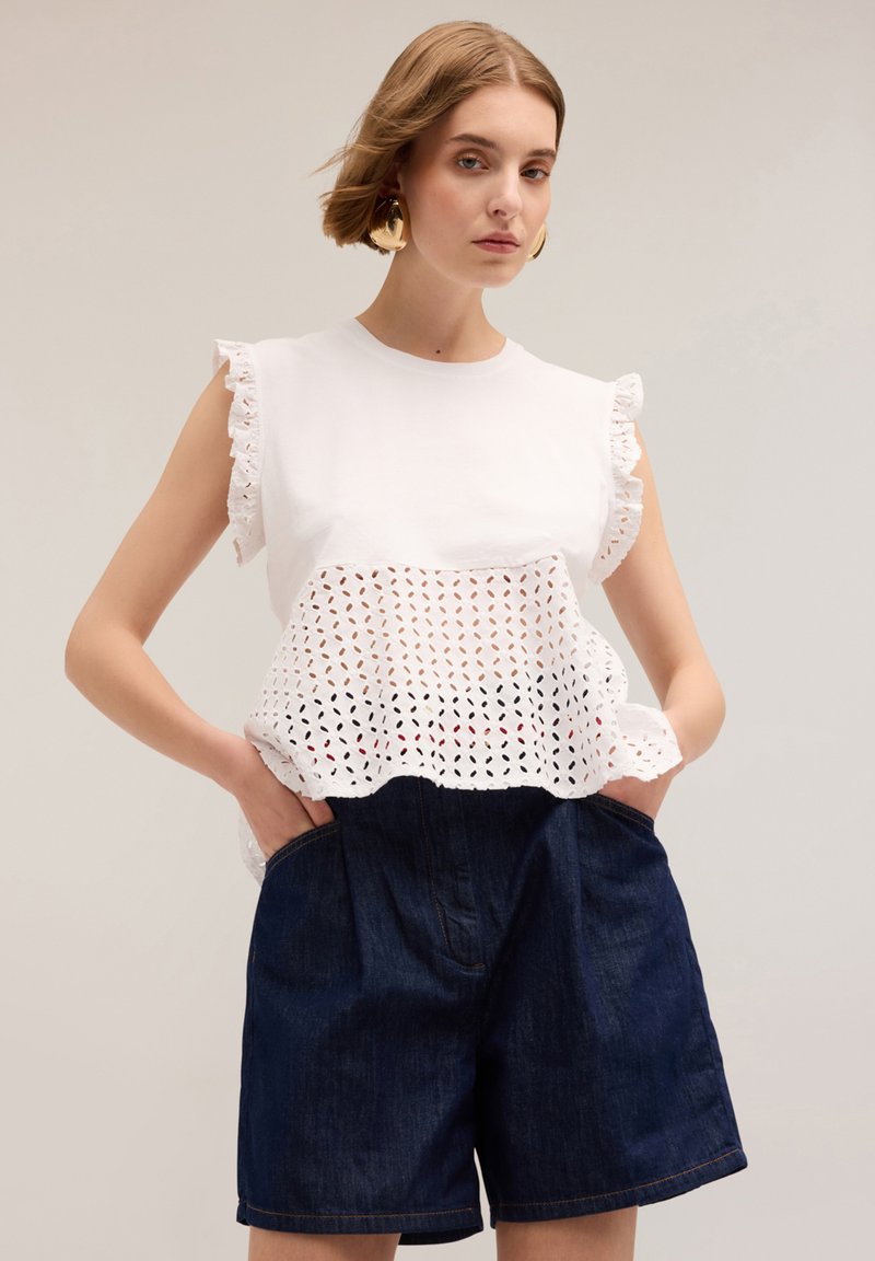 Top blanco con diseño de ojal y mangas con volantes, combinado con pantalones cortos de mezclilla azul oscuro que cuentan con bolsillos frontales y un ajuste relajado.