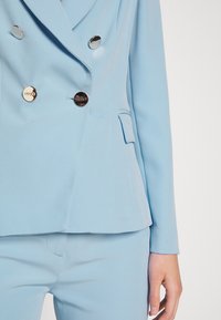 Jasnoniebieska dwurzędowa marynarka z gładkiego materiału, z srebrnymi okrągłymi guzikami i detalem bocznej kieszeni.