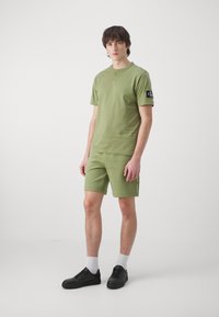 Camiseta de algodón verde oliva y pantalones cortos a juego, con un parche del logo de CK en la manga. Completado con zapatillas negras y calcetines blancos.