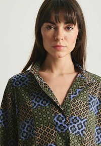 Chemise à col avec un design de patchwork présentant des motifs floraux et géométriques dans des teintes de vert, bleu et marron ; texture lisse.