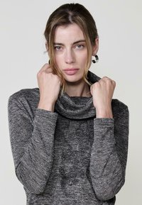 Grauer Cowl-Neck-Top aus strukturiertem Stoff, mit langen Ärmeln und einem dezenten floralen Muster auf der Vorderseite. Das Modell hält den Kragen.