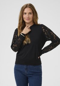 Jauna sieviete ar gariem matiem, valkājot melnu džemperi ar mežģīņu piedurknēm un zilos džinsos, pozē ar vienu roku pie sejas un otru uz gurniem.