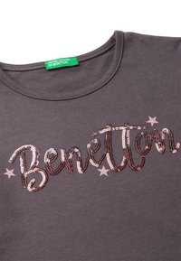 T-shirt in cotone grigia con collo rotondo presenta la parola "Benetton" in lettering sbrilluccicante e piccole stelle rosa. Etichetta verde sul collo.