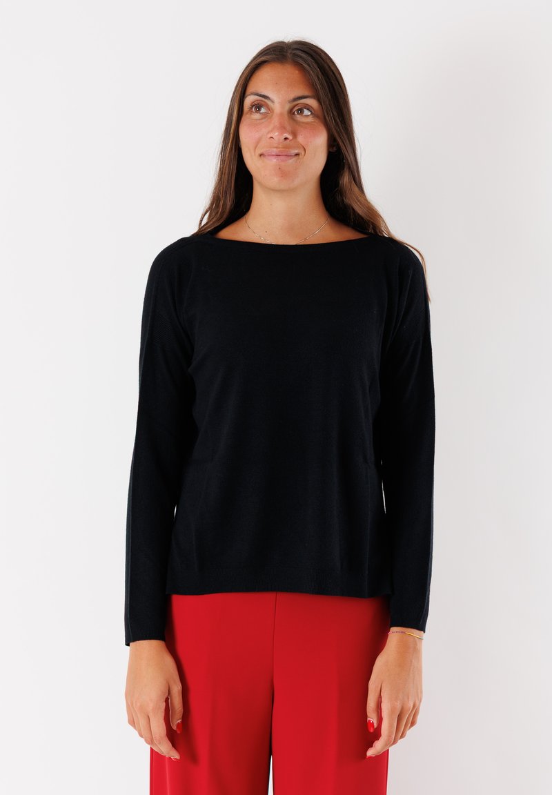 Maglione nero a maniche lunghe realizzato in tessuto morbido, con un ampio scollo e una vestibilità rilassata. Indossato con pantaloni rossi, creando un contrasto.