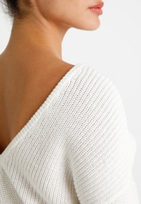 Valkoinen neulepaita, jossa on leveä V-pääntie, ja jonka materiaalissa on teksturoitu pinta ja ribbireunat kauluksessa ja hihoissa.