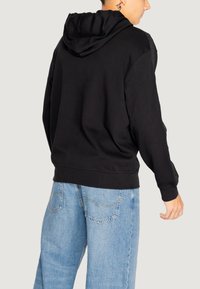 Felpa nera con cappuccio e vestibilità comoda, realizzata in tessuto morbido, con polsini a costine e dettagli laterali. Abbinata a jeans baggy blu chiaro.