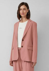 Vrouw in een stoffige roze blazer en bijpassende broek over een witte blouse met de tekst "S. Oliver", staande tegen een effen lichtgrijze achtergrond.