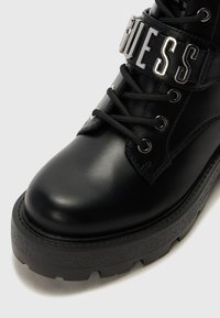 Sorte lær ankle boot med rund tå, kraftig sort såle, snøring foran og en metallrem med ordet "GUESS."