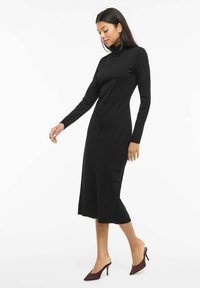 Černé šaty s dlouhým rukávem a rolákovým límcem, s přiléhavým střihem a délkou sahající ke kolenům. Kombinované s bordó lodičkami na vysokém podpatku.