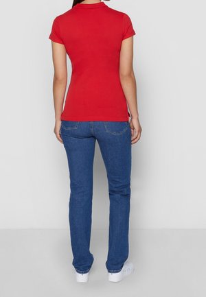 Kvinde stående med ryggen til iført en rød kortærmet polo, blå jeans og hvide sneakers mod en ensfarvet baggrund.