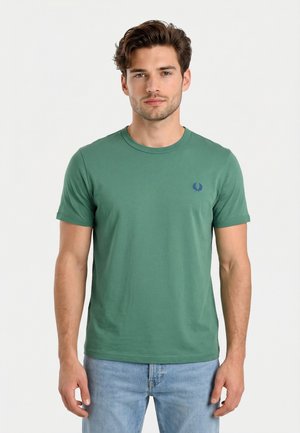 Giovane uomo che indossa una t-shirt verde a maniche corte con scollatura rotonda e jeans azzurri, in piedi davanti a uno sfondo bianco uniforme.
