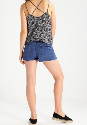 Zwarte bloemen tanktop met gekruiste banden, gecombineerd met blauwe denim shorts. Zwarte instapschoenen. Witte achtergrond. Casual outfit.