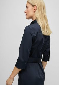 Sötétkék ruha övvel a derékban, háromnegyedes ujjal és galléros nyakkivágással. Simogatott anyag, strukturált sziluett.