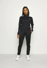 Chándal negro que incluye una chaqueta con cremallera y rayas blancas en las mangas, junto con pantalones a juego. Fabricado con una tela suave y elástica. Zapatillas blancas.