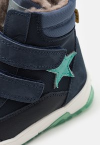 Mörkblå barnkänga med kardborreband, mjukt foder i päls och en ljusgrön stjärndetalj. Gummisula med en genomskinlig grön kant.