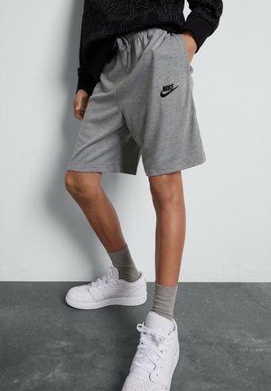 Personne portant un short Nike gris, des chaussettes grises, des baskets blanches et un sweat-shirt noir, debout contre un mur gris clair et un sol sombre.