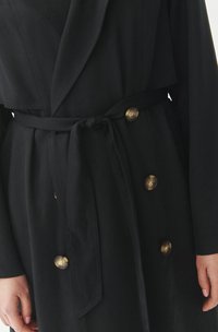Black coat with a shawl collar, double-breasted design, gold-tone buttons, and a tied waist belt. Made from a soft, textured fabric.