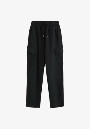 Teenager med krøllet hår iført en lilla t-shirt med grafik, sorte cargobukser, hvide sneakers og en sølvkæde, står foran en hvid baggrund.