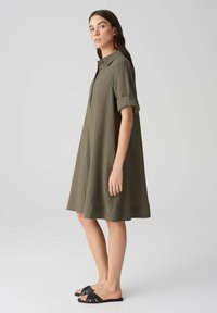 Vestito camicia verde oliva con colletto, maniche corte arrotolate, texture a pieghe e forma A-line svasata. Indossato con sandali neri a infradito.