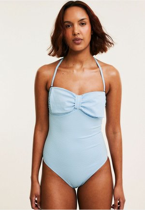 Frau mit schulterlangem braunen Haar, die einen hellblau-weiß gestreiften Einteiler mit Bandeau-Oberteil und Neckholder-Trägern trägt.