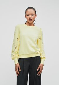 Myk gul genser med puffermer, har dekorative utskårne blomster på ermene, ribbet mansjetter og rund halslinning.