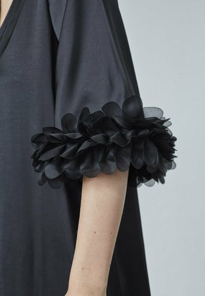 Sort beklædning med en dyb V-udskæring og voluminøse ærmer, der har lagdelte sorte stofblomster ved manchetterne, hvilket giver tekstur og designdetaljer.