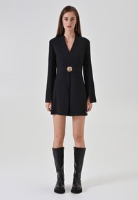 Donna che indossa un mini abito nero a maniche lunghe con una spilla d'oro a forma di fiore in vita e stivali neri fino al ginocchio su uno sfondo neutro.