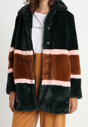 Vrouw draagt een donker groen, bruin en roze kleurvlakken kunstbontjas over een wit t-shirt en beige broek.