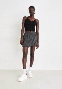 Czarny krótki top na ramiączkach z falbaniastymi brzegami, w połączeniu z szaro-paskowaną mini spódnicą i białymi trampkami z czerwonymi akcentami. Modelka stoi na neutralnym tle.