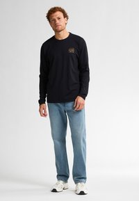 Schwarzes langärmliges T-Shirt mit einem kleinen Grafikdesign auf der Brust, kombiniert mit hellblauen Jeans und weißen Sneakers. Schlichte, lässige Gestaltung.