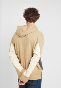 Sudadera con capucha beige con mangas en crema y gris. Presenta un ajuste holgado y puños acanalados. Tela de algodón suave con textura lisa.