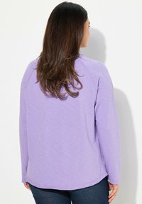 Felpa lunga viola con maniche lunghe e vestibilità rilassata, colletto a costine e orlo arrotondato. Tessuto morbido con una leggera texture, che mette in evidenza le cuciture sulle spalle.