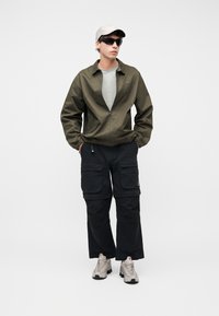 Νεαρός άνδρας που φοράει χακί μπουφάν, ανοιχτό γκρι καπέλο, μαύρα γυαλιά ηλίου, μαύρο παντελόνι καρό και μπεζ αθλητικά παπούτσια, στέκεται με τα χέρια στις τσέπες.