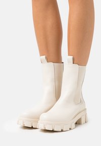 Bottines en cuir blanc avec une semelle épaisse, dotées de panneaux latéraux élastiques et de languettes pour un enfilage facile. Texture lisse et design à bout arrondi.