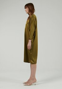 Vestido de satén verde oliva con un corte holgado, escote redondo y mangas tres cuartos, combinado con sandalias transparentes de tacón.