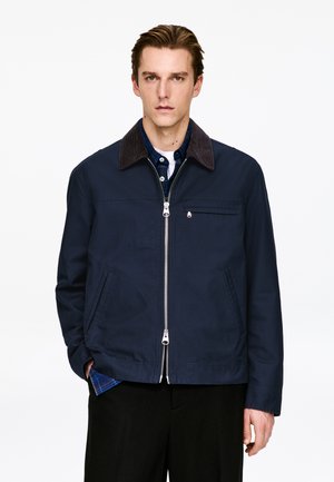 Granatowa kurtka z kołnierzem z sztruksu, zamkiem błyskawicznym z przodu, dwoma bocznymi kieszeniami oraz kieszenią na piersi. Charakteryzuje się gładką fakturą i minimalnymi detalami.