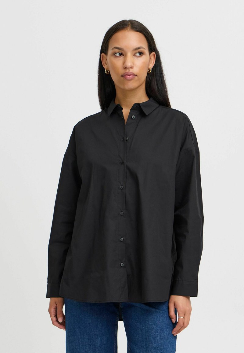 Camisa negra de botones con tejido suave, cuello tipo solapa, corte holgado y mangas largas. La parte frontal cuenta con botones negros y un diseño minimalista.