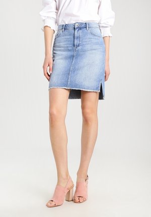 Kvinde iført en lyseblå denimnederdel med frontslids, hvid bluse og lyserøde åbne blokhælssandaler, stående på en ensfarvet baggrund.