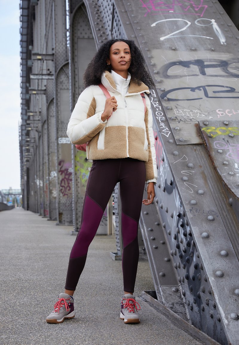 Giacca a vento con pannelli bianchi e beige, leggings neri e bordeaux aderenti, e sneakers grigie con lacci rossi, in piedi contro una struttura metallica.