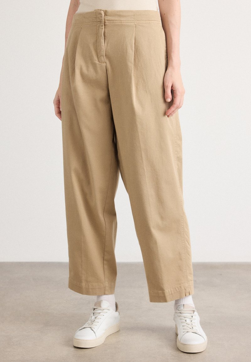 Beige wijde broek van katoen met een soepele textuur, met een hoge taille, plooien aan de voorkant en een iets verkorte lengte. Gecombineerd met witte sneakers.