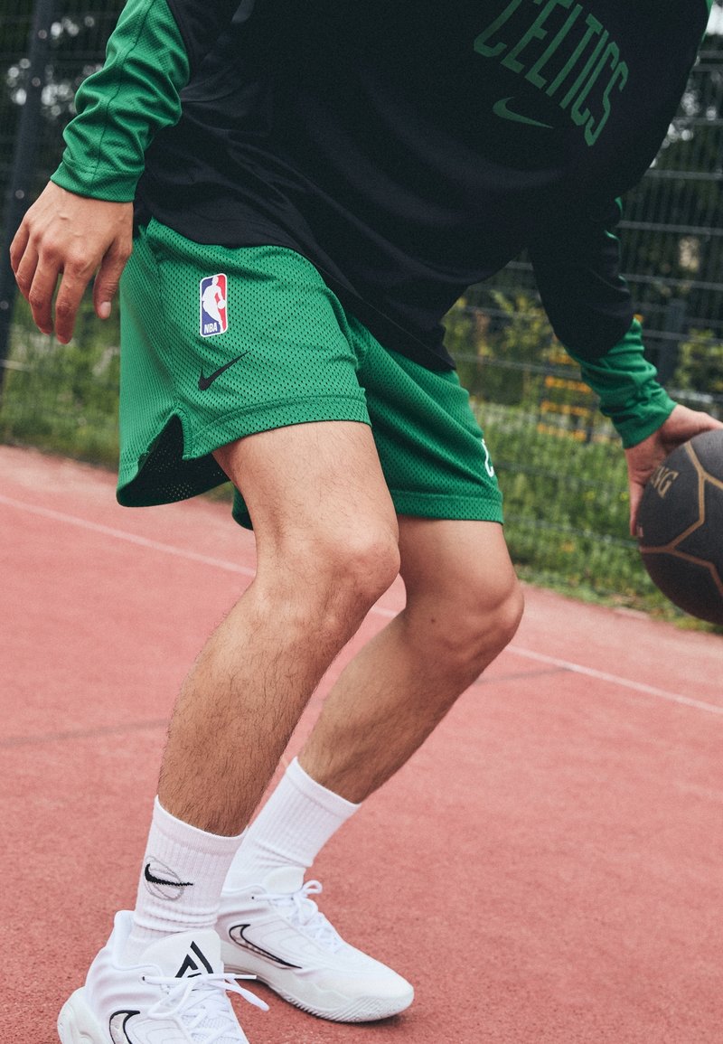 Gröna mesh basketshorts med NBA-logotyp, i kombination med en svart långärmad topp. Vita sportskor och ribbade strumpor på en rosa court.
