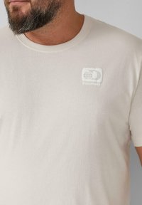 Cremfarbener Baumwoll-T-Shirt mit Rundhalsausschnitt, ausgestattet mit einem geprägten Discovery-Logo auf der linken Brust. Weicher Stoff, kurze Ärmeln.