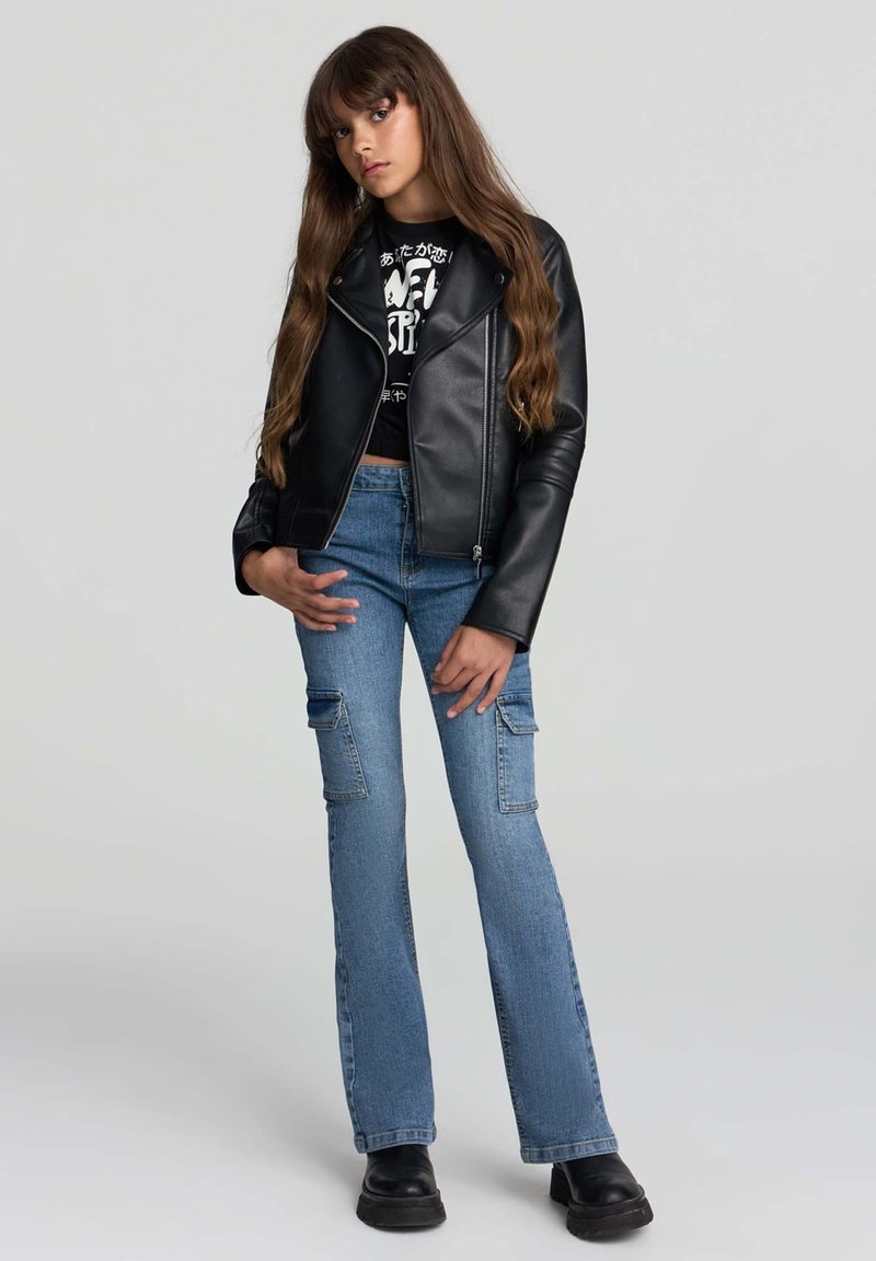 Ragazza adolescente con lunghi capelli castani, che indossa una giacca di pelle nera, un crop top grafico nero, jeans cargo blu e stivali neri su sfondo grigio.