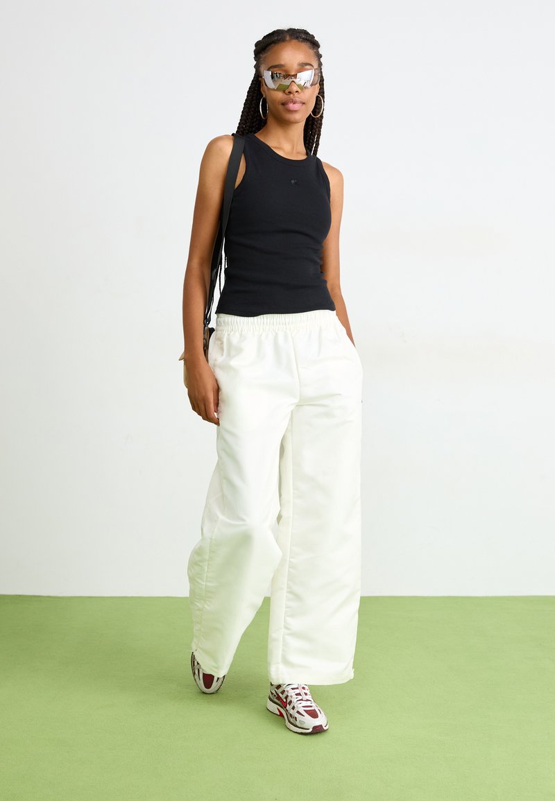 Un haut noir sans manches associé à un pantalon blanc ample. Le mannequin porte des lunettes de soleil miroir et des chaussures de sport avec des accents rouges.