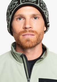 Zielona polarowa kurtka z wysokim kołnierzem, zamkiem błyskawicznym i czarnymi akcentami. Noszona z wzorzystą czarno-zieloną czapką na drutach.