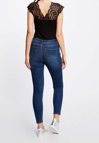 Žena v tmavě modrých skinny džínách a černém topu s průhlednou krajkovou zadní částí, v kombinaci se stříbrnými teniskami, stojící na jednobarevném pozadí.
