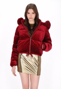 Chaqueta tipo puffer de terciopelo rojo con cierre de cremallera, dobladillo con cordón y pompones de piel en la capucha. Llevada sobre una minifalda dorada brillante.