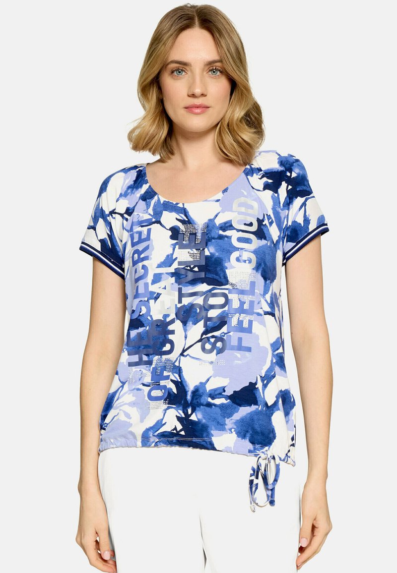 Femme portant un haut manches courtes bleu et blanc à imprimé floral avec un texte en strass et un détail lien à la base.