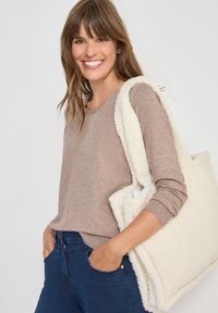 Hellbraunes Langarmshirt, kombiniert mit dunkelblauen Jeans. Die Tasche ist aus strukturiertem weißen Kunstfell mit dicken Trägern und einem geräumigen Design.