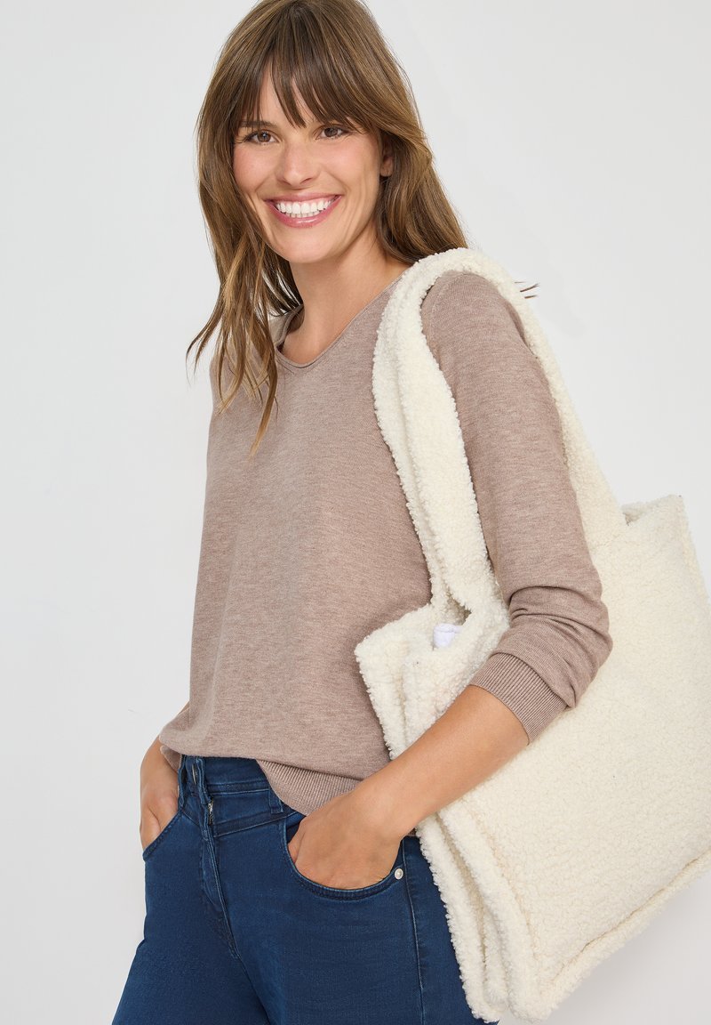 Hellbraunes Langarmshirt, kombiniert mit dunkelblauen Jeans. Die Tasche ist aus strukturiertem weißen Kunstfell mit dicken Trägern und einem geräumigen Design.